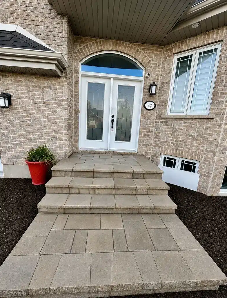 Escalier extérieur en béton rénové avec marches en dalles beiges, menant à une porte double blanche d’une maison en brique