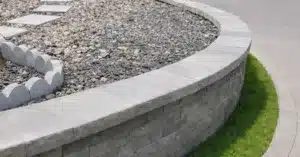 Mur de soutènement courbé en blocs de béton gris avec pierre de couronnement, bordé de gazon et d’un trottoir